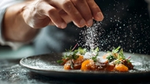 Chef sprinkling salt on gourmet dish with herbs on a dark plate.